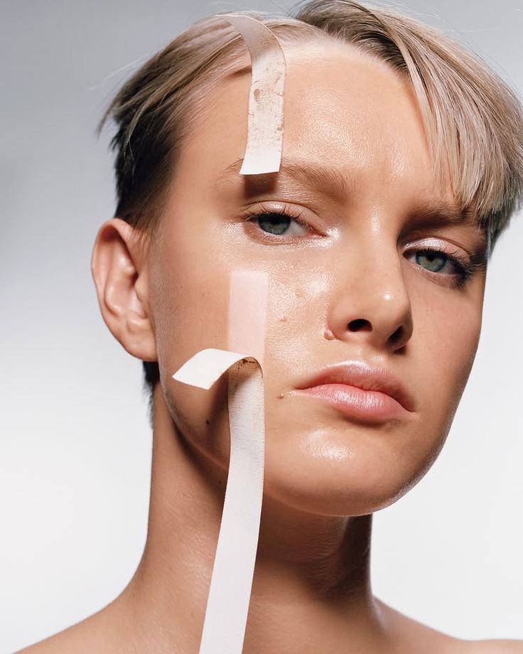 Close up portrait of a model with short blonde hair and glowing, hydrated skin, with strips of pale tape across the forehead and cheek, representing the skin barrier